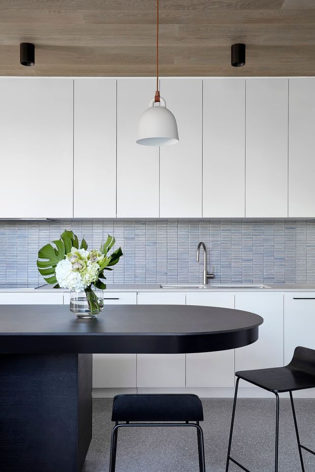 Contemporary minimalist kitchen featuring white full-height cabinetry, a horizontally patterned grey tiled splashback, dark curved island benchtop with integrated seating, black chairs, and warm timber ceiling panelling. A sculptural pendant light and black recessed spotlights provide focused illumination, while a floral arrangement adds a natural accent.