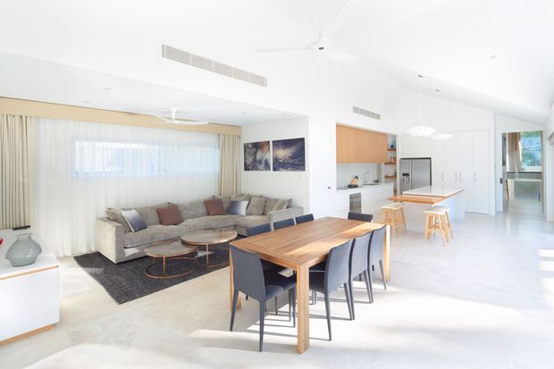 Bright open-plan contemporary home interior combining a living room, dining area, and kitchen. The space features white walls and ceilings, pale flooring, sheer curtains, a large neutral sectional sofa, a timber dining table with dark upholstered chairs, a timber kitchen island with stools, and abundant natural light.