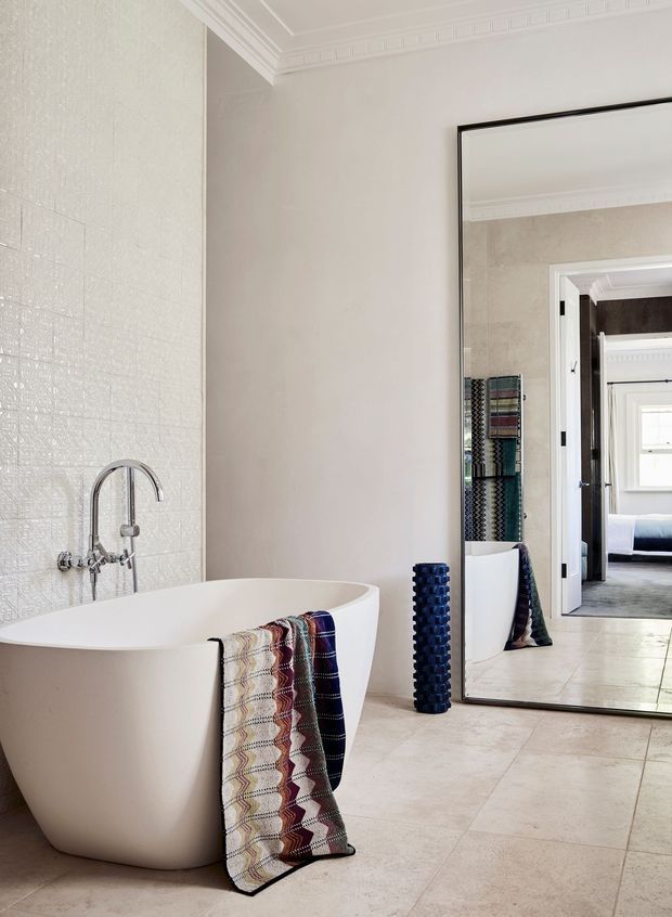 Elegant bright bathroom featuring a freestanding white bathtub beside a textured white tiled wall with a chrome wall-mounted faucet. Large pale stone-look floor tiles, ornate white ceiling cornice, and a full-height black-framed mirror create a refined traditional setting. The mirror reflects an adjoining doorway and bedroom beyond. A patterned textile hangs over the tub, with a dark blue decorative vessel on the floor.
