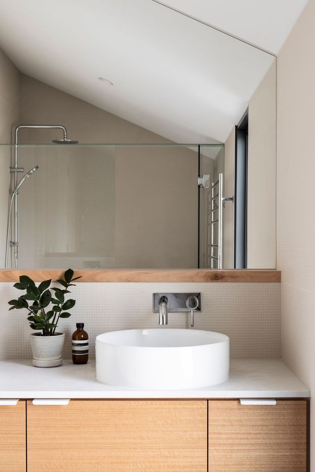 A contemporary bathroom vanity with a white vessel basin, wall-mounted chrome tapware, pale timber cabinetry, and a warm timber shelf beneath a large frameless mirror. A glass-enclosed shower with chrome fixtures is visible behind the vanity. The room has beige textured wall tiles, a light neutral benchtop, and a sloped white ceiling, creating a clean, restrained and softly warm interior.