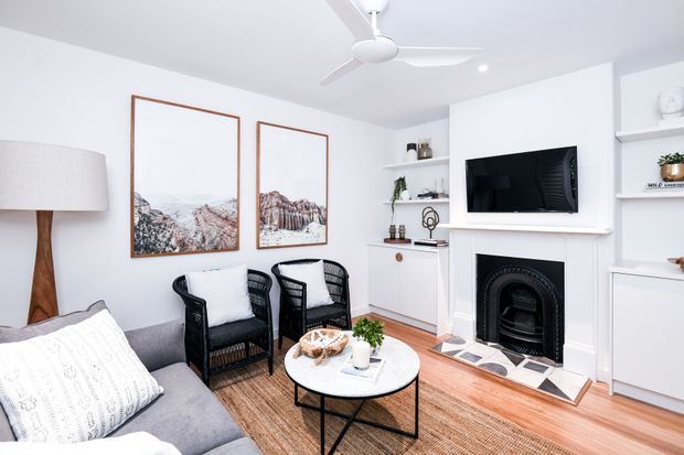 Bright, compact living room with white built-in shelving and cabinetry, a wall-mounted television above a traditional black fireplace, grey sofa, black woven armchairs, round coffee table, timber flooring, and framed artwork. A white ceiling fan and recessed lighting complete the clean, refined interior.