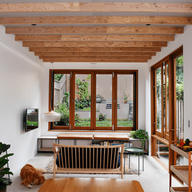 Bright, narrow living room with exposed timber ceiling beams, white walls, warm timber-framed windows and glazed doors, and a large garden-facing window. The room includes a mid-century-style slatted sofa, low timber coffee table, bench seating beneath the window, indoor plants, a television, and a small dining or serving area at the right. The space feels warm, natural, and inviting.