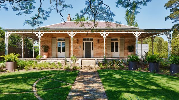 Front exterior view of a single-storey heritage-style timber cottage with a long verandah, decorative white timber fretwork, multiple entry doors, a low hipped roof, and a landscaped garden with a brick-paved path leading to the porch.