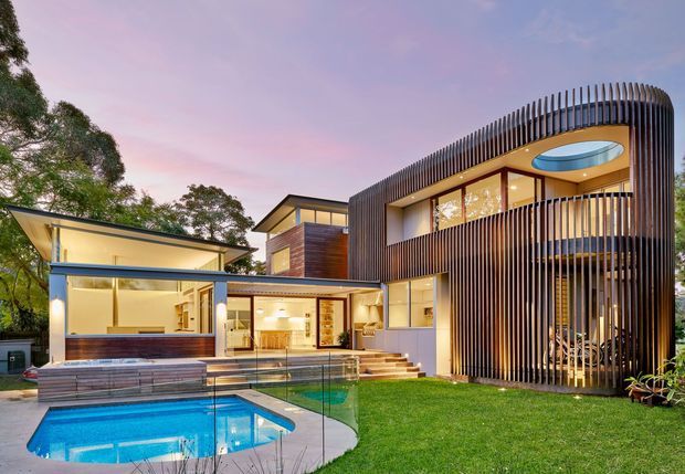 Contemporary multi-level residential home viewed from the landscaped rear garden, featuring extensive glazing, warm timber cladding, a curved vertical timber-screen facade, and a partially enclosed swimming pool terrace. The upper level includes a distinctive oval rooflight or skylight, while the lower level opens toward the pool and lawn. The image is captured at dusk with warm interior lighting and a pink-purple sky.
