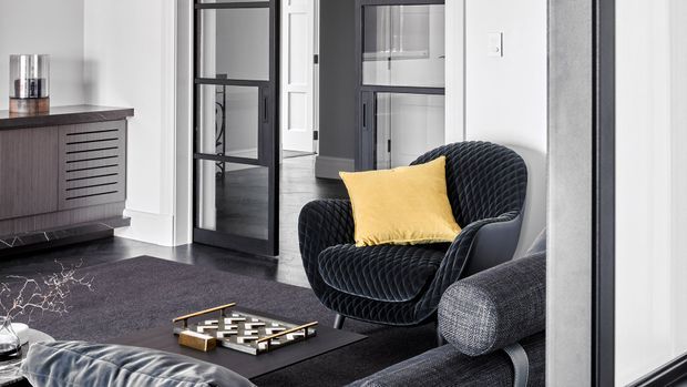 A sophisticated modern living room featuring a dark textured rug, sculptural black lounge chair with a mustard-yellow cushion, grey upholstered sofa, and dark timber sideboard with slatted drawer fronts. Black-framed glazed internal doors and crisp white walls create a strong monochrome architectural backdrop, with contemporary artwork and accessories adding subtle detail.