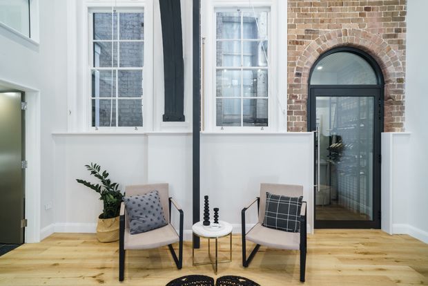 Bright, refined sitting area within a renovated brick building, featuring white wall panelling, tall multi-pane windows, a black-framed arched glazed doorway, and warm timber flooring. Two contemporary lounge chairs flank a small round side table, with minimal plant and cushion accents.
