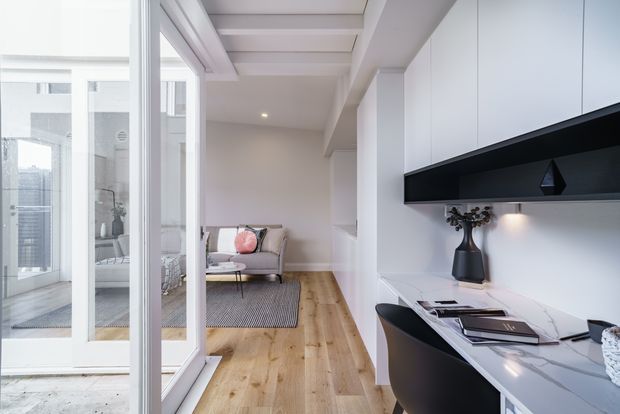 Bright contemporary open-plan interior viewed from a compact kitchen or study nook toward a living room. The space features white built-in cabinetry, a dark linear overhead feature, a pale stone work surface, light timber flooring, glass-panelled doors, exposed ceiling beams, and a small upholstered sofa in the adjoining room.