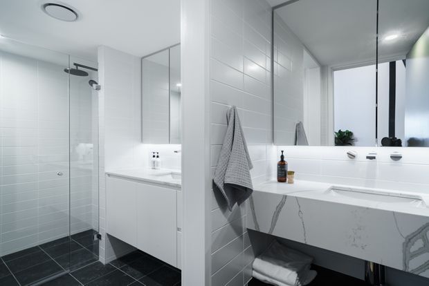 Bright contemporary bathroom with a walk-in glass shower on the left and a freestanding sculptural bathtub on the right. The room features white horizontal wall tiles, dark charcoal floor tiles, a white floating vanity with integrated basin, large mirrors, frameless shower glazing, and minimal chrome fixtures. A narrow vertical window brings in natural light.