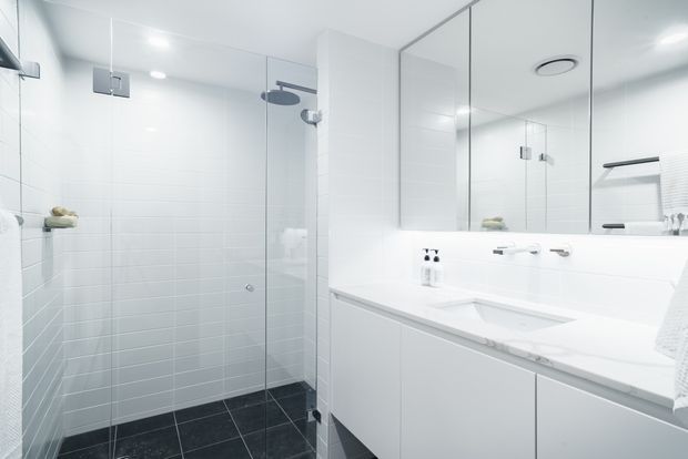 Bright, clean contemporary bathroom with a frameless glass walk-in shower, white tiled walls, dark tiled floor, and a long white vanity with integrated basin and mirrored wall cabinetry. Chrome fixtures and recessed lighting create a crisp, minimalist appearance.