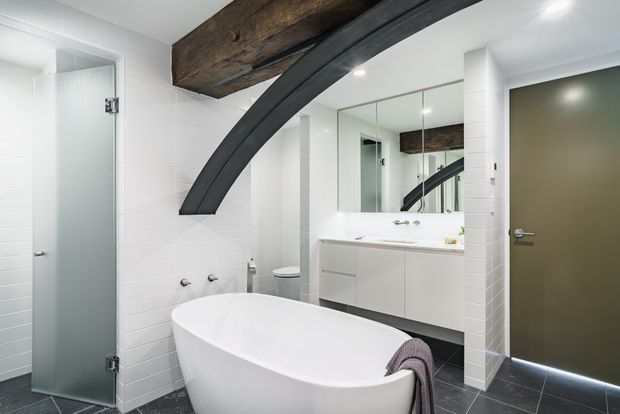 Contemporary bathroom with a freestanding white bathtub, glass-enclosed shower, wall-hung vanity with double mirrored cabinet, white tiled walls, dark floor tiles, and a dramatic exposed timber beam with a curved dark structural element across the ceiling.