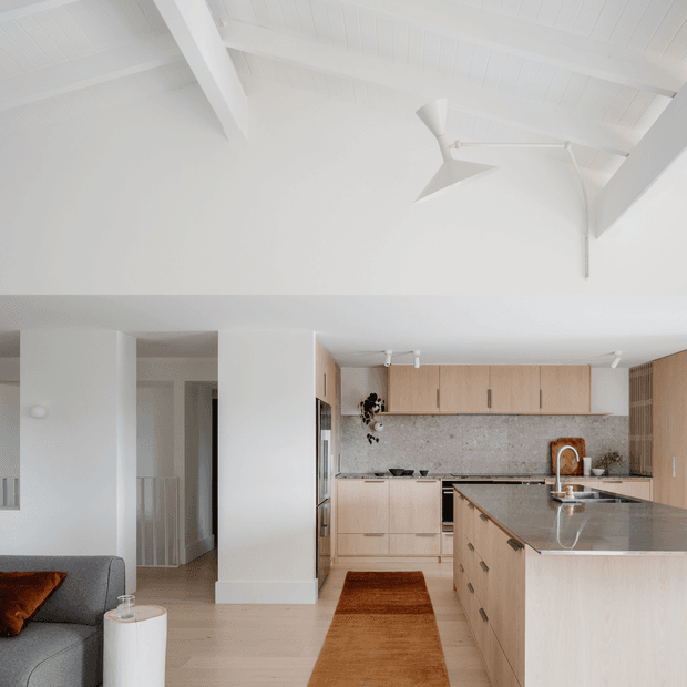 Bright open-plan kitchen and living area with a minimalist white interior, exposed white-painted ceiling beams, and a pitched ceiling. The kitchen features light timber cabinetry, a speckled stone splashback, a large stone island with integrated sink, and a modern angled ceiling fan. A grey sofa and rust-toned rug and cushion add warmth.