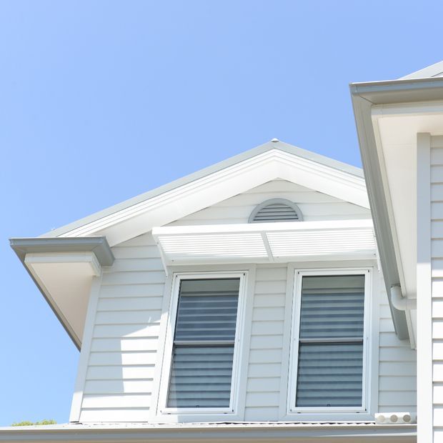 Close-up exterior view of a bright white weatherboard-clad residential facade, featuring a centered gable roof, decorative louvre vent, two tall windows with external blinds, and a projecting white awning. The composition highlights traditional detailing against a clear blue sky.