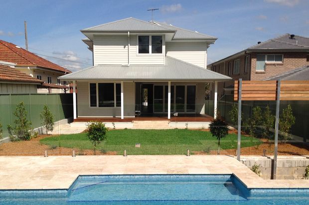 Street-facing view of a two-storey residential house with pale weatherboard cladding, a light grey hipped metal roof, covered front veranda, landscaped lawn, paved forecourt, and a swimming pool in the foreground. The home sits among neighbouring suburban houses.