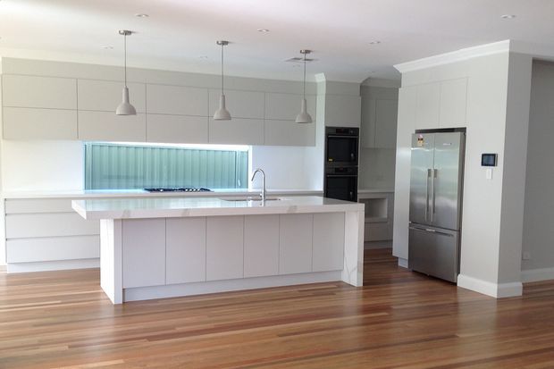 Bright contemporary kitchen with a large white island, integrated cabinetry, stainless-steel appliances, pendant lighting, and warm timber flooring. A vertical ribbed glass splashback or window feature sits behind the cooktop, with a dining or living area opening to the right.