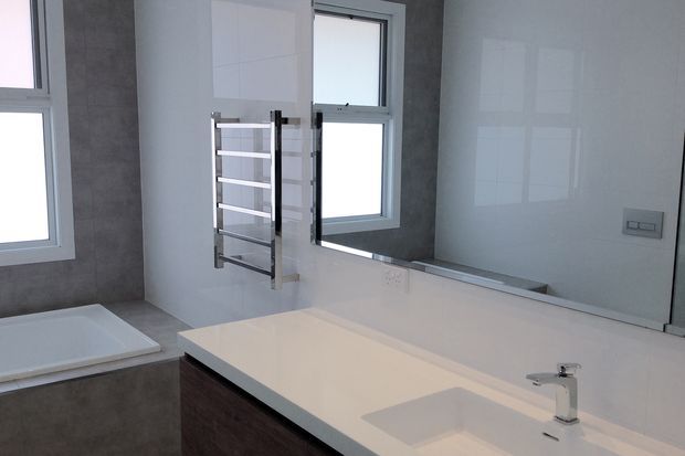 A bright contemporary bathroom with a large wall-to-wall mirror, white tiled walls, grey textured wall tiles, a white countertop with integrated basin and chrome tap, dark wood-toned vanity cabinetry, a wall-mounted heated towel rail, and a partially visible bathtub beneath a window.