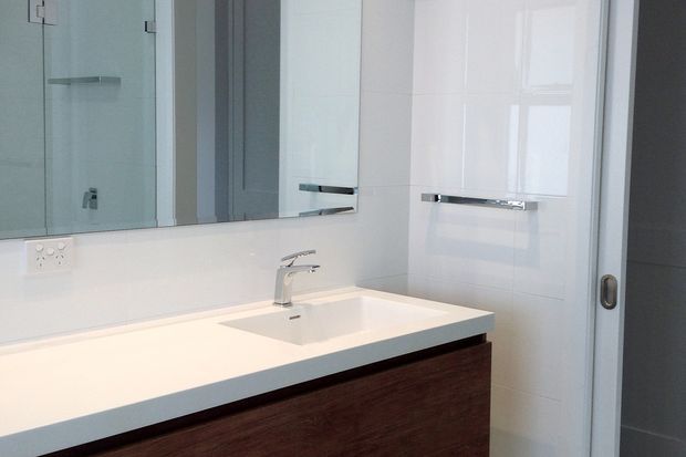 Bright, compact contemporary bathroom vanity with a white integrated basin and benchtop, dark timber-look cabinetry, chrome tapware and handles, large wall mirror, and a glazed shower enclosure.