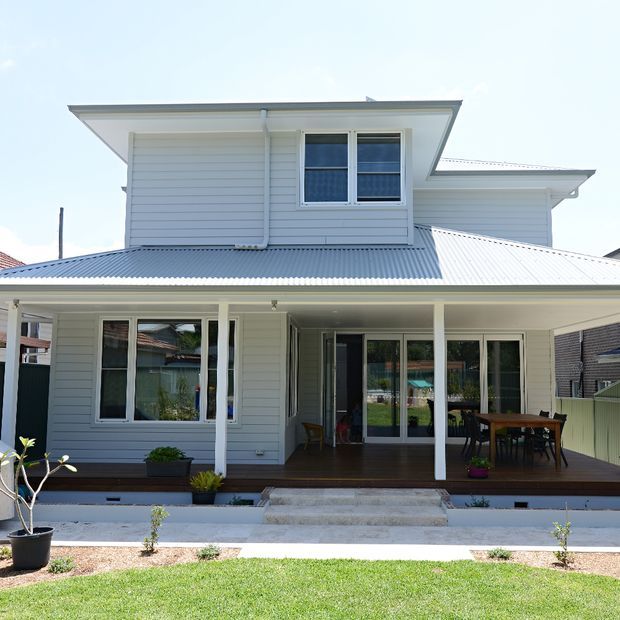 Front exterior view of a two-storey suburban house with white horizontal weatherboard cladding, a broad corrugated metal roof, and a covered ground-floor veranda/deck. White-framed windows and glazed doors open onto an outdoor dining area, with simple landscaped planting in the foreground.