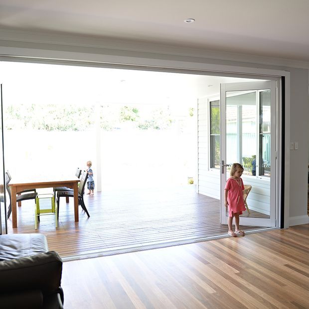 Bright contemporary residential interior viewed from a living room through wide-open glazed doors to a covered timber deck or sunroom. The foreground has warm timber flooring and a dark sofa, while the adjoining space contains a timber dining table and chairs, white wall cladding, and extensive glazing.