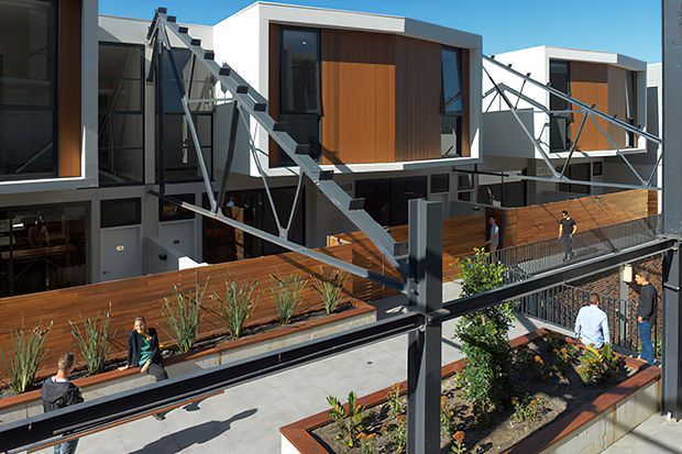 Exterior view of a contemporary multi-level residential development with box-like white volumes, dark-framed glazing, warm timber cladding, and prominent exposed steel bracing. Elevated pedestrian walkways, private terraces, planter beds, and landscaped communal areas connect the buildings.