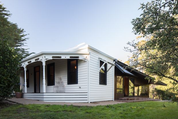 Exterior view of a small white weatherboard cottage with a front verandah, decorative timber posts and fretwork, dark-trimmed windows, and a pitched metal roof. A glazed timber extension projects from the side, with the house set within a leafy garden.