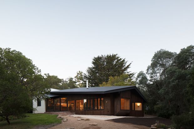 Single-storey dark timber-clad house set within a leafy landscaped site, photographed from the front at dusk. The low-pitched dark roof, broad glazed living-area frontage, projecting timber volume, and warm interior lighting create a restrained contemporary cabin-like appearance.
