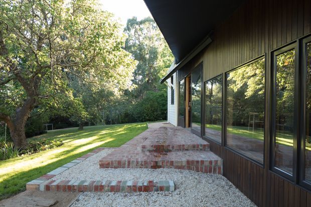 Exterior view of a low-slung house opening onto a landscaped garden. The facade combines dark vertical timber cladding with large dark-framed glazed doors and windows, while broad brick-edged steps lead across a pale gravel terrace. Mature trees, lawn, and reflected greenery create a secluded garden setting.