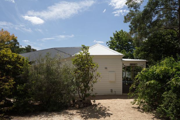 Exterior view of a small, single-storey light-coloured weatherboard building set within mature garden landscaping. The building has a simple gable roof with pale metal roofing, a covered entry or side verandah, and a gravel or pale stone pathway leading through dense shrubs and trees. Bright daylight and blue sky create a relaxed residential garden setting.