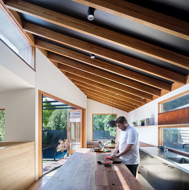 Bright contemporary kitchen with a long central timber island, integrated stainless-steel cabinetry and appliances, extensive timber-framed glazing, and a dramatic exposed timber-beam ceiling against dark painted recesses. The open-plan space connects visually to an outdoor area through the glazed door and windows.