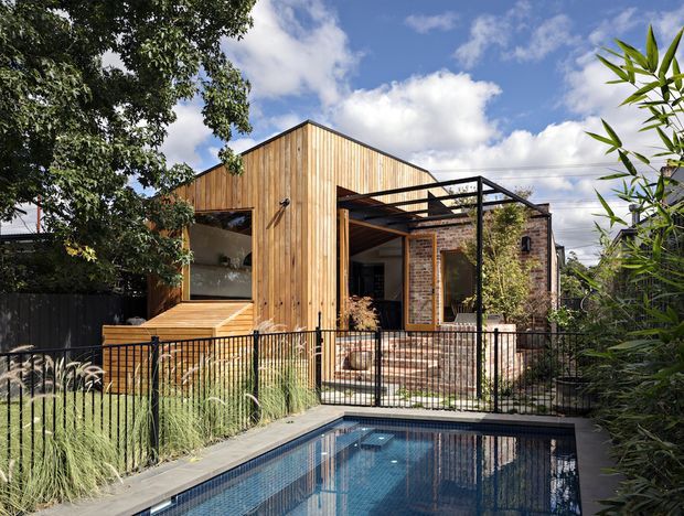 Exterior view of a contemporary residential extension with a steeply pitched gable form clad in vertical timber boards, adjoining exposed brick walls and a black steel pergola. A timber deck, landscaped garden, metal fencing and a narrow swimming pool occupy the foreground.