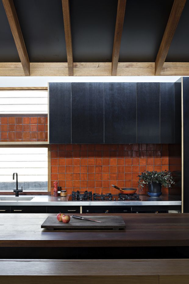 A dramatic, material-rich kitchen interior with a long dark timber island in the foreground, black fixed cabinetry, stainless-steel benchtops, and a vivid orange-red tiled splashback. A gas cooktop, sink, tapware, cookware, and potted plant are visible. Large horizontal windows bring in natural light, while exposed dark timber roof rafters and a black ceiling create a strong architectural overhead feature.