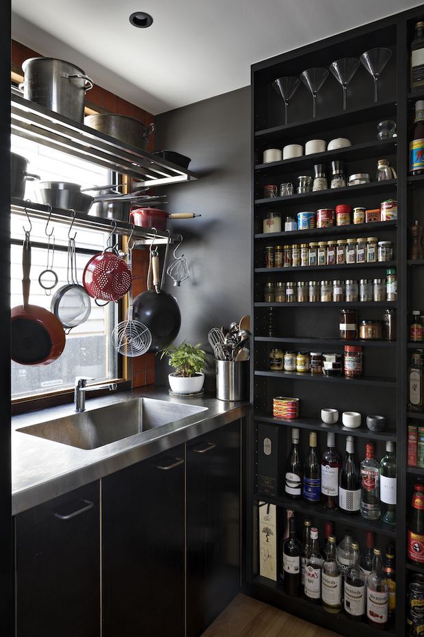 A compact, highly organised kitchen with a stainless-steel sink and benchtop, black lower cabinetry, open black shelving filled with glassware, spices, pantry goods and bottles, and extensive hanging cookware beneath a metal overhead rack. A window provides strong natural light, while dark finishes and exposed storage create a practical, utilitarian character.