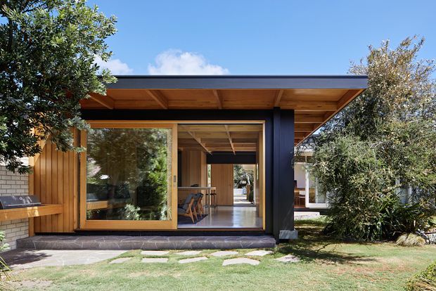 Single-storey modern pavilion-style house extension viewed from the garden, featuring a broad flat roof with exposed timber soffit, extensive timber-framed glazing, vertical timber cladding, and a dark structural wall or column. The open glazed frontage reveals a timber-lined interior and a long axial view through the dwelling. A paved path and landscaped lawn sit in the foreground.