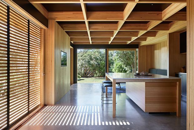 A warm, timber-lined open-plan kitchen and dining space with a sculptural exposed timber ceiling grid, a large central island with bar stools, full-height glazing to a leafy garden, and timber louvred screening casting strong striped shadows across the polished concrete floor.