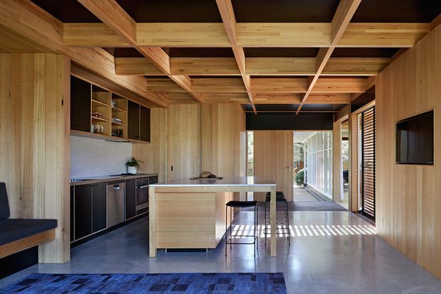A contemporary open-plan kitchen and living area with extensive warm timber wall panelling, cabinetry, island bench and exposed coffered ceiling beams. The kitchen has dark lower cabinetry, a pale benchtop and integrated appliances, while a long dining table extends from the island. Concrete flooring, strong linear daylight through louvred glazing, and a wall-mounted television create a refined indoor-outdoor atmosphere.