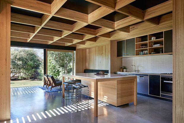 Open-plan kitchen and dining area with a large timber island, stools, dark built-in cabinetry, integrated shelving, and a tiled backsplash. The room features extensive warm timber wall panelling and a distinctive exposed timber coffered ceiling, with large glazed doors opening toward a leafy outdoor area. Strong sunlight creates striped shadows across the polished concrete floor.