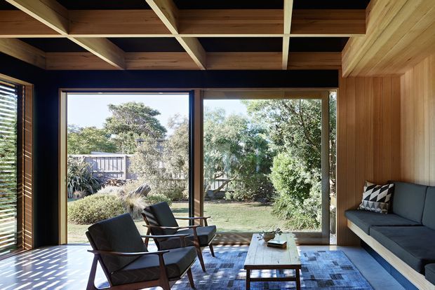 A warm, mid-century-inspired living room with full-height glazing opening onto a landscaped garden. The room features a built-in dark upholstered sofa, two timber-and-leather lounge chairs, a low timber coffee table, richly toned timber wall cladding, and a distinctive exposed timber ceiling grid with dark recessed panels. Strong daylight and garden views create an indoor–outdoor connection.