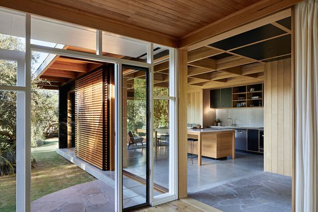 A warm, timber-lined interior looking through full-height glazed doors toward a compact contemporary kitchen and adjoining outdoor space. The room features exposed timber ceiling beams and wall framing, dark cabinetry, a pale tiled backsplash, timber island cabinetry, and a dark stone-look floor. Exterior timber screening and garden views are visible through the open glazing.