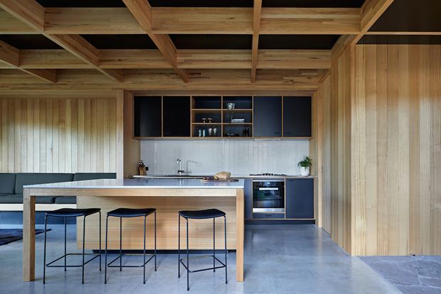 Open-plan contemporary kitchen and dining area with a long timber island and three slim black bar stools. The room features extensive warm timber wall panelling, exposed timber ceiling beams with dark infill panels, dark blue-black cabinetry, a pale textured backsplash, and a polished grey concrete floor. A built-in oven and cooktop are integrated into the cabinetry, while a low upholstered seating area is visible at left.