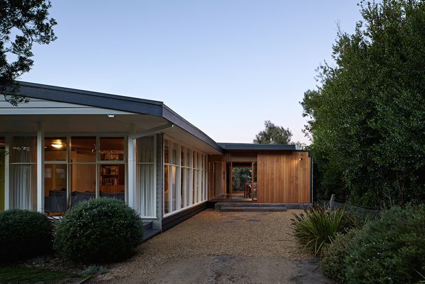 Single-storey modern residential house viewed from a landscaped gravel courtyard. The home has broad floor-to-ceiling glazing with slender dark frames, a low-pitched roof with pronounced eaves, and a warm timber-clad projecting volume at the rear. Interior furnishings are visible through the glazing, while mature planting frames the entry approach.