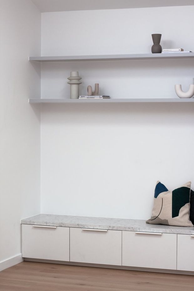 Minimalist built-in storage and display wall with two long floating shelves, neutral decorative objects, and low white cabinetry topped with a pale stone surface. The room has white walls, warm timber flooring, and restrained sculptural decor with a dark accent cushion.