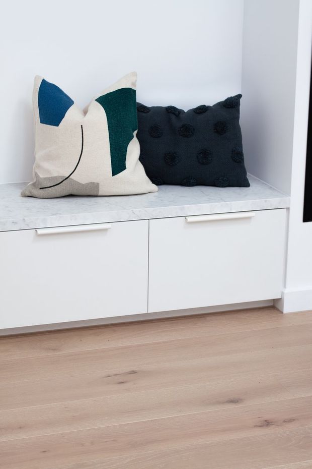A bright minimalist interior detail showing a low white built-in cabinet with slim horizontal handles and a pale marble-look benchtop. Two decorative cushions sit on the surface, one abstract cream, blue and green cushion and one dark navy textured cushion. Pale natural timber flooring and white walls create a clean, restrained palette.