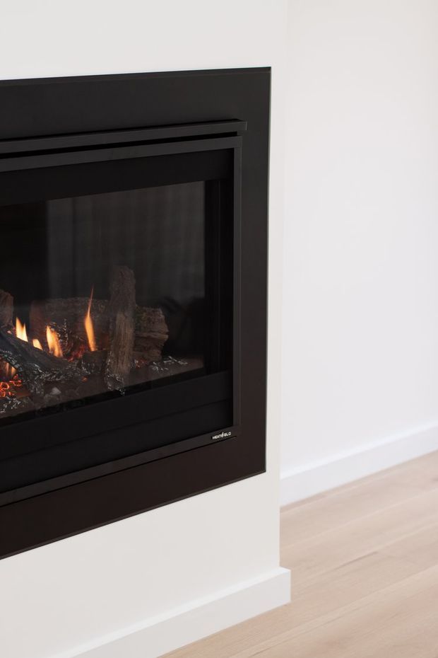 Close-up of a sleek built-in black fireplace with visible flames and log-effect interior, set into a smooth white wall. Pale timber flooring and simple white skirting create a clean, minimal interior.