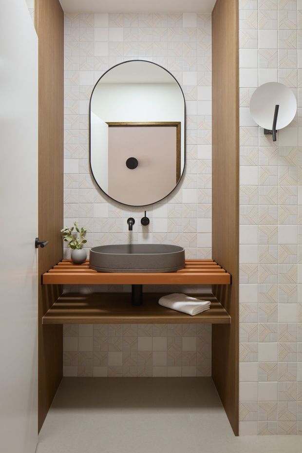 A compact contemporary bathroom vanity viewed straight on. The space features a wall-mounted oval vessel basin in dark grey, black tapware, a large rounded-rectangle mirror, patterned pale geometric wall tiles, warm timber side panels and open timber shelving. A circular wall light is visible on the right, with minimal accessories and a folded towel on the lower shelf.