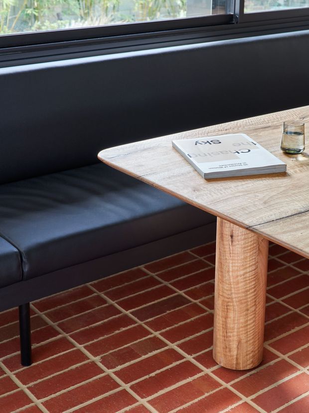 Close-up interior view of a contemporary seating nook with a black upholstered built-in bench beside a black-framed window. A light timber table with a rounded edge and substantial cylindrical timber leg sits over a red brick-tile floor with pale grout. A book and drinking glass rest on the table, while greenery is visible outside the window.