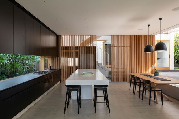 A spacious contemporary open-plan kitchen and dining area with dark cabinetry along the left wall, a long pale island with integrated seating, and a timber dining table on the right. Vertical timber screening and slatted feature walls define the central stair and circulation zone, while large windows and indoor planting bring in natural light and greenery. Black pendant lights and furniture provide strong contrast against the warm timber and neutral concrete-toned surfaces.