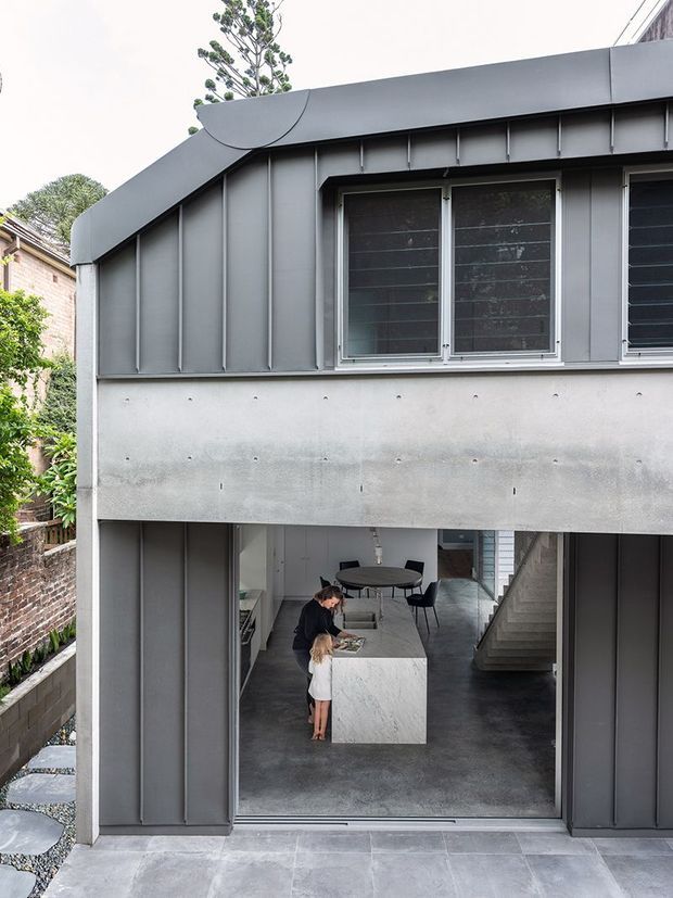 Exterior view of a contemporary two-storey home with a strong grey metal-clad upper facade, projecting concrete band, and large opening revealing an open-plan kitchen and dining area. The lower level has a dark polished concrete floor, a pale stone kitchen island, black dining furniture, and a visible internal stair. A narrow landscaped side passage and neighbouring masonry walls are visible.