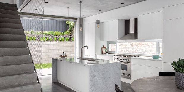 Contemporary open-plan kitchen with white flat-panel cabinetry, a large pale stone island, stainless-steel appliances, and a concrete ceiling. A dark carpeted staircase rises alongside the kitchen, while full-height glazing opens to a planted courtyard. A round timber dining table is visible in the foreground.