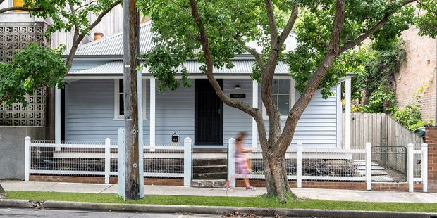 Street-facing view of a compact single-storey residential home with pale horizontal weatherboard cladding, a corrugated metal pitched roof, dark-framed windows and entry door, and a small fenced front garden. Mature street trees partially obscure the facade, while decorative breeze-block screening and neighbouring masonry walls appear at the sides.