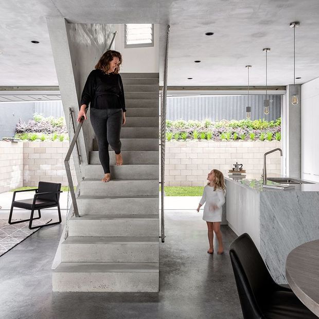 Contemporary open-plan ground-floor interior with a central exposed concrete staircase, polished concrete flooring, and a minimalist kitchen. The kitchen features a large grey stone island with an integrated sink, tall curved tap, pendant lights, and dark dining chairs. Full-height glazing opens toward a small landscaped courtyard, while a child and adult are visible in the space. The raw concrete finishes and clean-lined detailing create an industrial-modern atmosphere.
