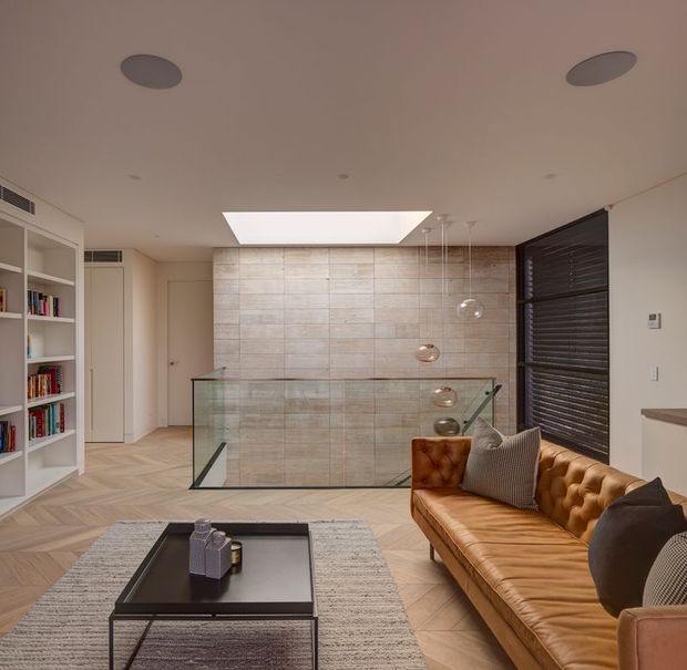 A refined contemporary living room overlooking a lower-level stairwell or void through a clear glass balustrade. The room features a caramel leather button-tufted sofa, black rectangular coffee table, textured rug, built-in white shelving filled with books, pale herringbone timber flooring, a tiled feature wall, recessed lighting, a large skylight, and dark horizontal window blinds.